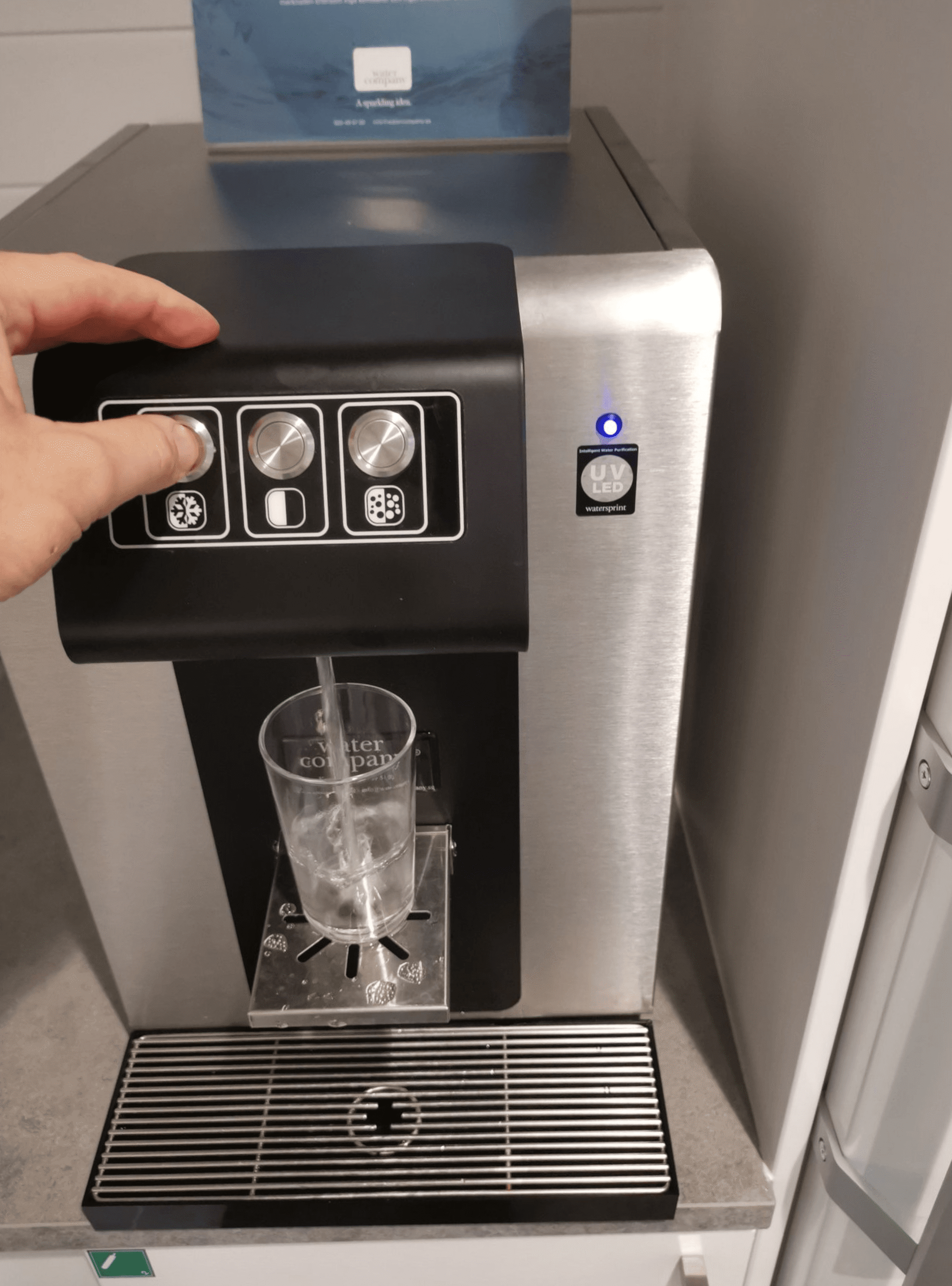 En som fyller på ett vattenglas från en vattenautomat som står på en bänk.