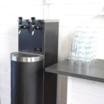 Water Companys vattenautomat Tower i svart med två vattenkranar för kolsyra och stilla. Vattenautomaten står bredvid en bänk med vattenglas.