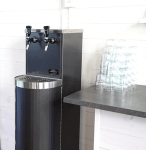 Water Companys vattenautomat Tower i svart med två vattenkranar för kolsyra och stilla. Vattenautomaten står bredvid en bänk med vattenglas.