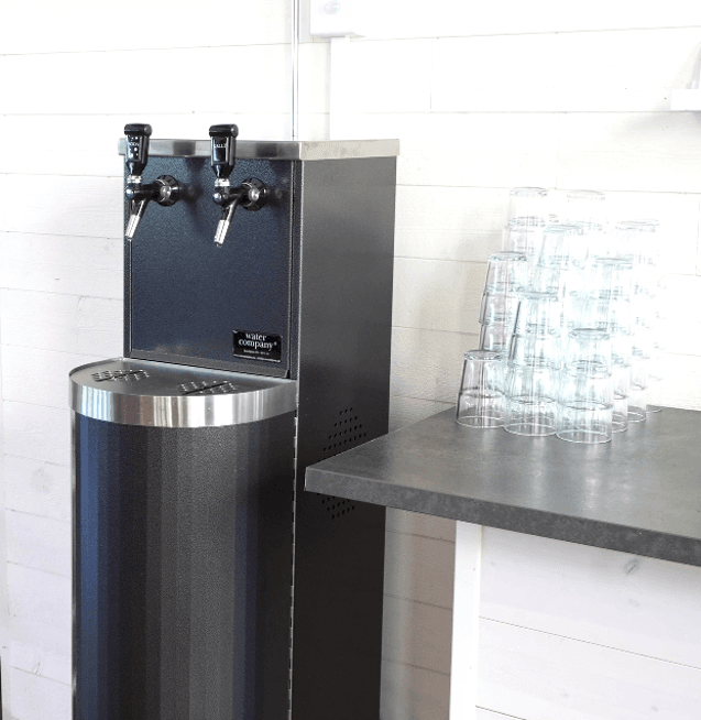Water Companys vattenautomat Tower i svart med två vattenkranar för kolsyra och stilla. Vattenautomaten står bredvid en bänk med vattenglas.