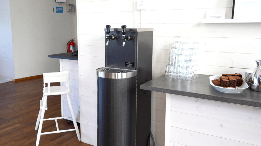 Water Companys vattenautomat Tower i svart med två vattenkranar för kolsyra och stilla. Vattenautomaten står bredvid en bänk med vattenglas.