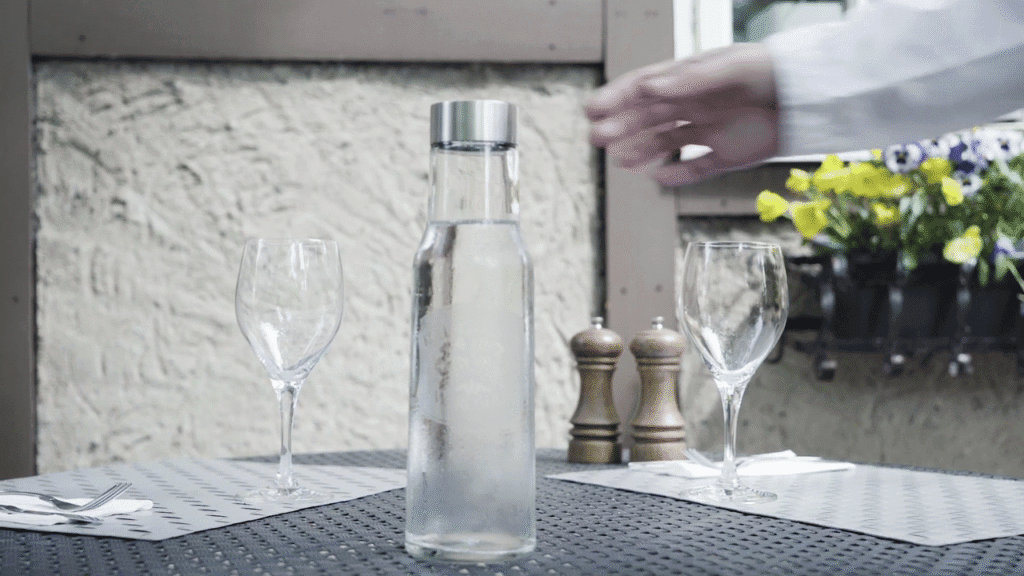 Ett dukat bord med två fyllda vattenglas och en vattenkaraff i mitten gjort av glas med graverad logotyp.